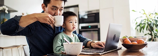 A father checks his laptop while drinking a cup of coffee with his son on his lap.
